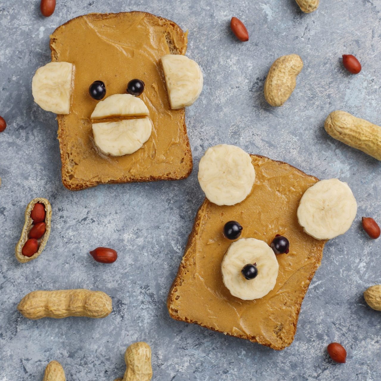 Funny bear and monkey face sandwich with peanut butter, banana and black currant,peanuts on grey concrete background,top view